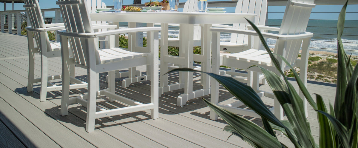 A white Trex Outdoor Furniture Cape Cod Adirondack 7 Piece Dining Set sits on a deck overlooking a beach and the ocean.