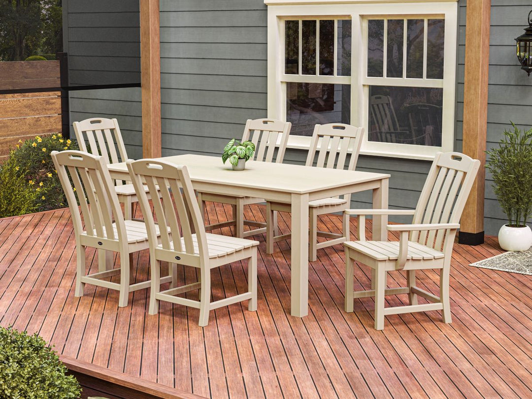 A tan Trex Outdoor Furniture Yacht Club 7 Piece Parsons Dining Set sits on a wooden deck by a green house with white windows.