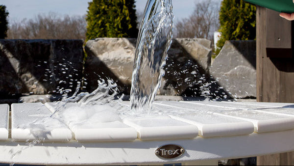 Water is being poured from a green bucket onto a round white Trex Outdoor Furniture table.